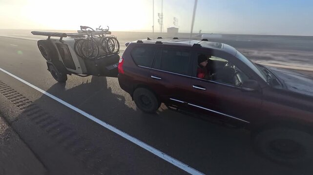 A mature woman sits in an SUV looking out at the view while towing a trailer with bicycles on a long road.