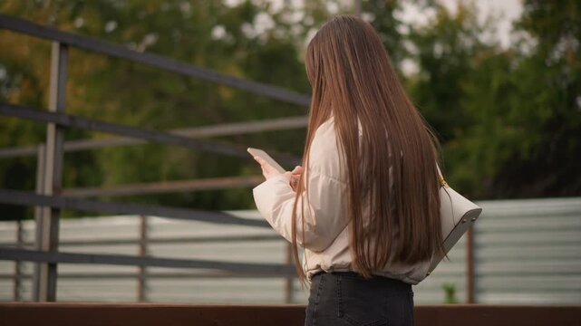 Caucasian woman long hair checking phone near metal railing, beige jacket and tote bag, overcast sky and autumn foliage in background, slow handheld pan captures candid scrolling and typing, subtle
