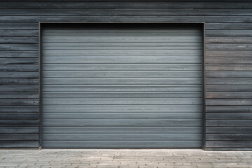 a closed gray roller shutter door on a dark wooden wall beside a stone pavement
