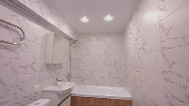 Modern bathroom with marble-patterned tiles, white sink, toilet, bathtub with wooden cabinet, heated towel rail, mirror above sink, clean minimalist design