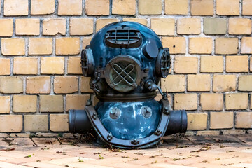 Photograph of an old and antique metal diving helmet sitting on the ground outside near a brick wall in the sunshine.