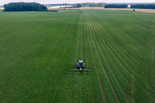 Aerial view of a lone tractor cutting hay into even windrows on European farmland, bordered by compact forest stands under diffuse daylight, emphasizing precision and order.