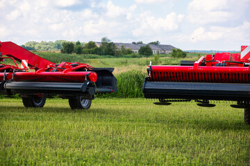 Two red forage harvester headers with black pickup reels stand on short grass, wheels and hydraulic arms visible. A low farm building and tree line sit in the distance.