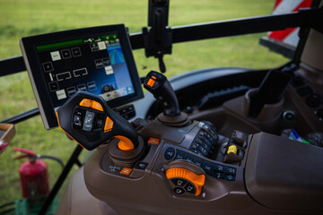 Close up of a modern tractor cockpit shows a touchscreen guidance display, joystick with labeled buttons A and B, numeric controls, rotary dials, and hydraulic levers in daylight.