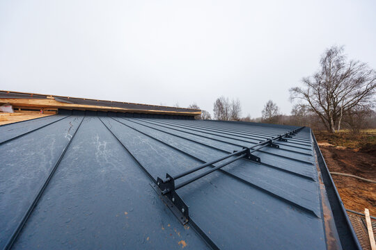 Low angle view of dark standing seam roof under construction, double rail snow guard spans panels, exposed eaves and underlayment at left, birch trees, wet panels, overcast light.