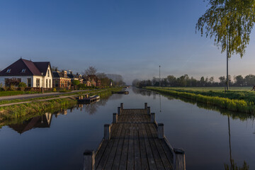 Obraz premium morning view from a wooden pier over the bedum canal. dutch landscape with mist, traditional houses and perfect reflection on the water