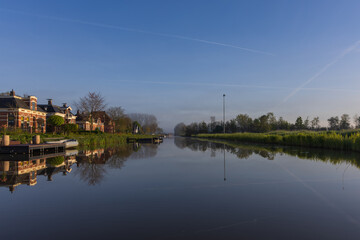 Fototapeta premium Peaceful morning canal in Bedum, Netherlands. Perfect reflections of houses and trees, morning haze on the horizon and clear blue sky sun