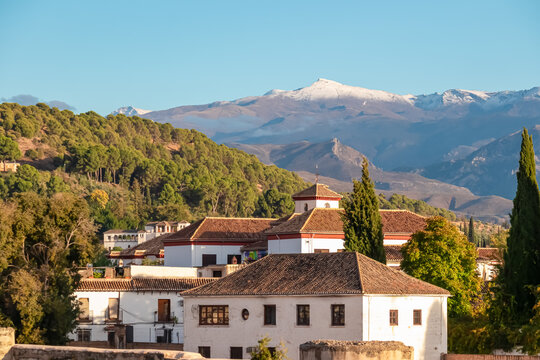 White traditional houses with tiled roofs stand against the snowy Sierra Nevada mountains in Granada, Spain, highlighting the contrast between the sunny city architecture and alpine peaks.