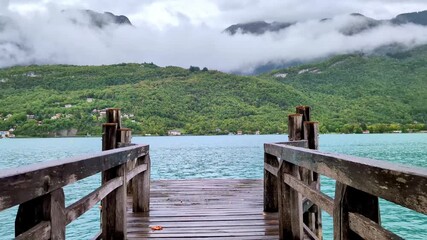 Scenic wooden pier on Lake Annecy extends over tranquil turquoise waters, framed by lush green Alps mountains and cloudy skies, showcasing nature's beauty of Talloires, France. - Powered by Adobe