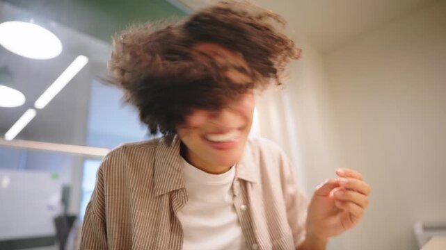 Happy young woman raising fists and smiling during video call, bright modern office interior with soft daylight showing success, excitement and positive remote communication moment