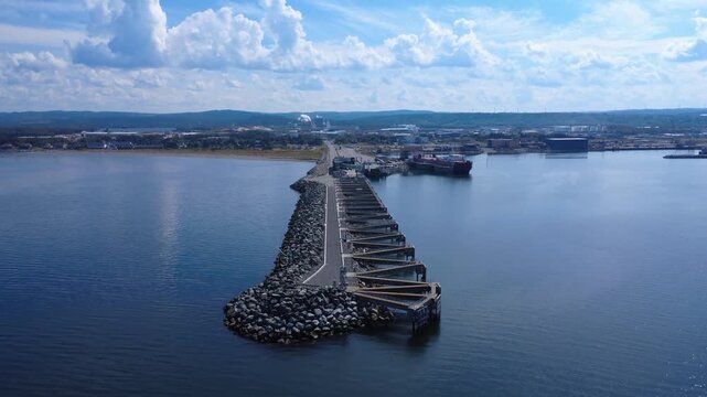 A long stone jetty juts out into a tranquil bay, connecting the shore to a bustling port. In the distance, warehouses and ships add an industrial touch to this peaceful seascape. Matane Port, Quebec.