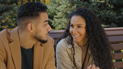 Talking positive Arabian Hispanic couple in city autumn park enjoying relationship date communication talk woman with man friendly talk conversation outdoors married girl with guy sitting on bench