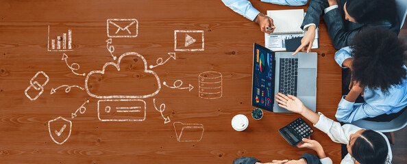 A diverse business team engages in a collaborative meeting focused on cloud computing. Digital graphics illustrate concepts on a wooden desk, enhancing creativity and strategy. Trope