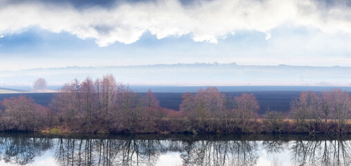 Autumn landscape with tree reflections in calm lake water under cloudy sky, natural panorama with fog and mirror water surface