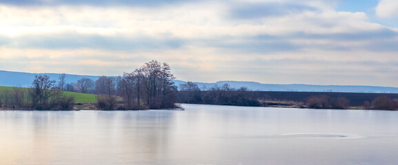Panoramic view of calm lake with island covered by trees and distant hills under soft cloudy sky in morning light