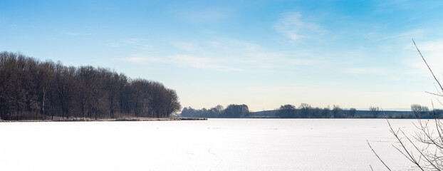Panoramic winter landscape with frozen lake covered by snow and forest on shore under blue sky with light clouds