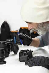 A bearded technician wearing gloves cleans a camera sensor using a specialized spatula, working carefully at a table surrounded by lenses and professional photography equipment.