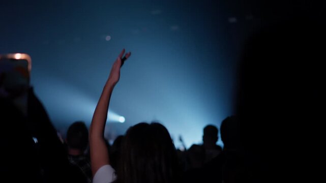 Silhouetted arm extends upward amid dense crowd bathed in blue spotlights highlighting energetic atmosphere at live music performance venue