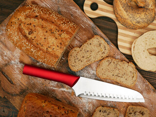 Various types of bread and bagels on wooden cutting boards, with a red-handled serrated knife. Some bread slices are also visible.