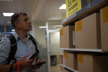 Man with backpack checking labeled cardboard boxes on shelf in Spanish postal office