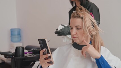 Caucasian woman in salon checking phone client sits in cape while stylist applies bleach and foils to braided hair, gloved hands adjust sections smartphone used for messages and mirror reference