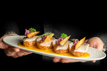 A close-up horizontal studio shot of gourmet herring appetizers on round toasted bread with pickled red onions, sliced pear, and dill on an oval white plate held by hands