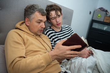 A father in a tan hoodie and son in a striped shirt read a book together while sitting on a bed at home