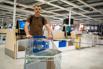 Man in brown shirt with backpack pushes a shopping cart in a large retail store aisle under bright lighting