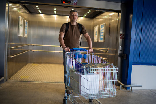 Man exiting an elevator while pushing a shopping cart loaded with items wearing a casual brown shirt and backpack indoors