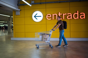 Man pushing a shopping cart with boxes in a parking area near a yellow wall with an entrance sign
