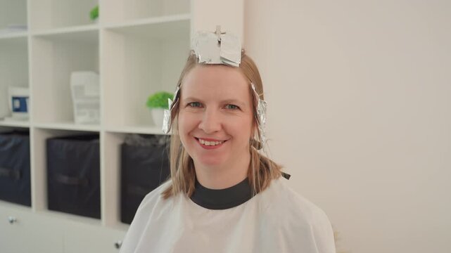smiling woman in salon with foil, beaming as stylist checks color and adjusts foils, protective cape worn, modern shelving and potted plant in background, bright warm lighting, candid joyful makeover