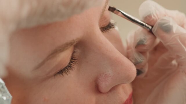 Closeup caucasian woman eyebrow microblading procedure, esthetician with gloved hands maps brow and applies pigment with precision tool, client relaxed with eyes closed, soft neutral lighting, sterile