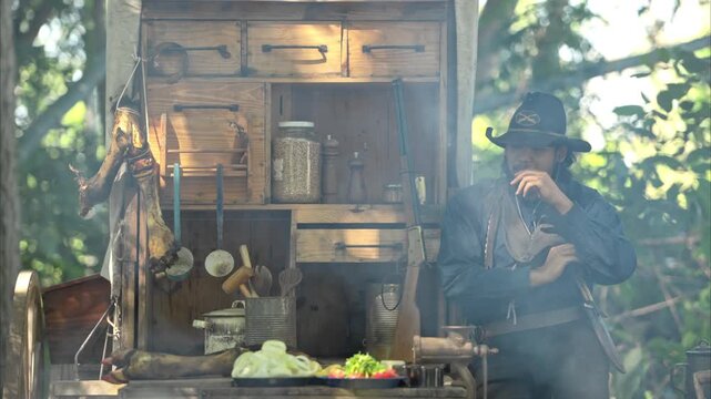 A cowboy stands holding a rifle in a forest camp, with sunlight streaming through the smoke and a chuck wagon in the background