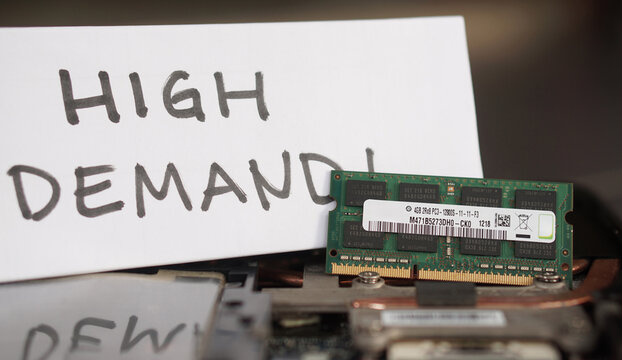 A DDR3 RAM stick resting on a laptop with a handwritten "HIGH DEMAND!" sign. Represents the global scarcity of electronic parts and supply chain bottlenecks.