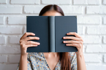 Woman sitting indoors holding a closed book covering her face against a white brick wall. Generative AI