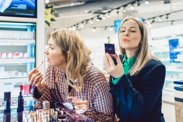 Two cheerful friends are choosing mascara and lipstick in cosmetics store. High quality photo