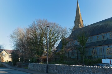 Stone church with tall spire dominates, flanked by bare trees, under bright blue sky; sunny, peaceful, distant street view, naturalistic photography in Caernarfon - Gwynedd - Wales - UK