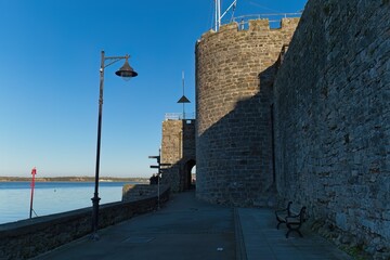 Stone castle wall and tower, shadowed path, blue water, clear sky, bright sunlight, lamppost, bench, creating a serene, historical, and slightly imposing atmosphere in Caernarfon - Gwynedd - Wales 