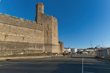 Imposing stone castle wall with a square tower dominates, bathed in bright sunlight under a clear blue sky, overlooking a paved area in Caernarfon - Gwynedd - Wales - UK