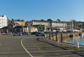 Harborfront townscape: colorful buildings line a marina with parked cars, under bright sunlight, creating a tranquil, picturesque scene with clear, reflective water in Caernarfon - Gwynedd - Wales 