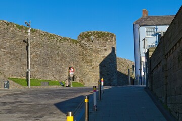 Sunlit stone city wall with a round tower dominates, adjacent to a blue building, under a clear sky, creating a historic, peaceful ambiance in Caernarfon - Gwynedd - Wales - UK
