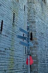 A stone castle wall looms behind a signpost with blank directional signs; gray stone, muted light, vertical composition, medieval ambiance, and slightly distant perspective in Caernarfon - uk