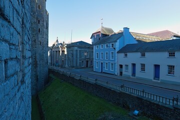 Stone castle wall overlooks a street with blue and white buildings under bright sunlight, creating a serene, historical ambiance with sharp architectural details in Caernarfon - Gwynedd - Wales - UK