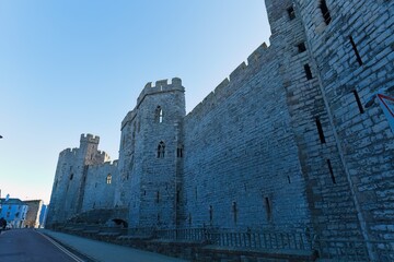 Imposing gray stone castle walls rise diagonally against a clear blue sky, featuring crenellations, narrow windows, and a somber, monumental atmosphere in Caernarfon - Gwynedd - Wales - UK