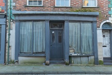 Abandoned storefront: grey facade, large windows with curtains, central door, brick building backdrop, overcast light, aged, melancholic, street-level view in Caernarfon - Gwynedd - Wales - UK