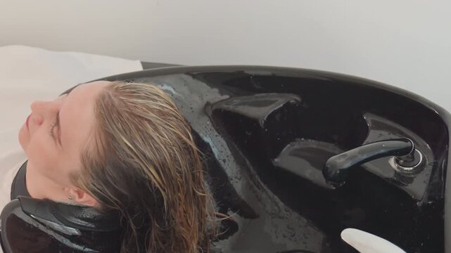 woman leaning over black basin as stylist adjusts faucet and rinses dyed roots with cool water, wet blonde strands, minimal modern interior, focused hands and clinical salon finish