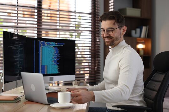 Smiling programmer in glasses working on computer with dual monitors and laptop at table in office - Powered by Adobe