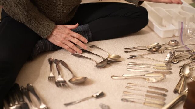 Mature woman sorts family heirloom silver cutlery on the floor of her home, carefully organizing each piece with focus and care.