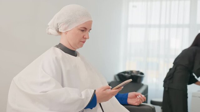 Caucasian client in cape checking phone, seated under processing cap, stylist preparing products in background, soft window light, minimalist white interior, calm patient mood during precolor