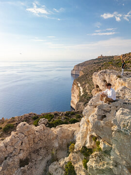 Man sitting on the edge of high yellow limestone Dingli Cliffs at sunset, scenic aerial view of Mediterranean sea and rocky coastline, Malta.
